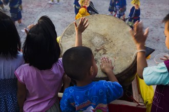 Thai children bang drum while watching dance troupe with firecrackers exploding at local temple.