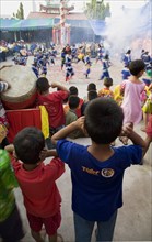 Thai children bang drum while watching dance troupe with firecrackers exploding at local temple.