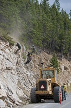 Workers wearing safety helmets on ropes clearing a rock slide and making the rock face safe along the Akamina Parkway Caterpillar tractor on parkway
