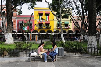Mexico, Puebla, Two young women chatting on stone bench in the Zocalo with colourful building