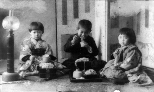 Three Japanese children seated on cushions on the floor, c.1900