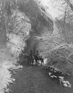 Two Crow Indians on horseback in shallow stream flanked by snow-covered trees