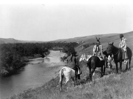 Three Piegan Indians and four horses on hill above river
