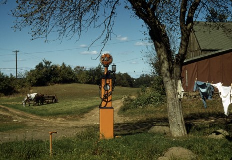 Gas pump with clothesline, barn and horse-drawn wagon in background.