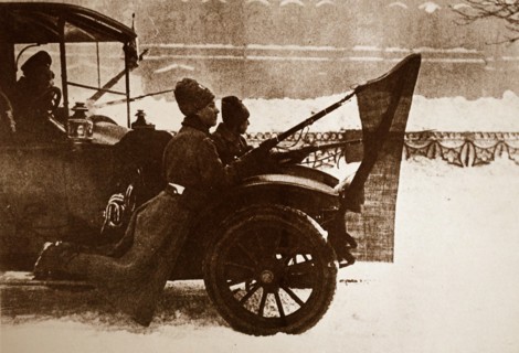 Photograph of soldiers riding on the mud flaps of a car with red flags attached to their guns in Petrograd