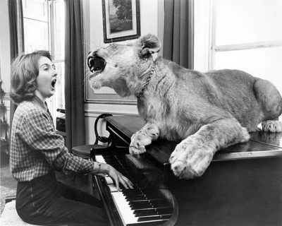 Lowe sits on a piano and sings together with the piano player