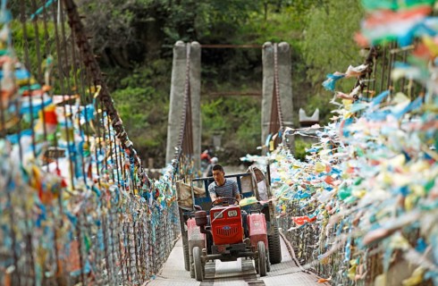Farmer drives with a tractor over a suspension bridge decorated with prayer flags