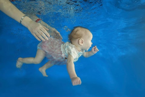 Infant girl dives in a pool with hands of her mother holding it