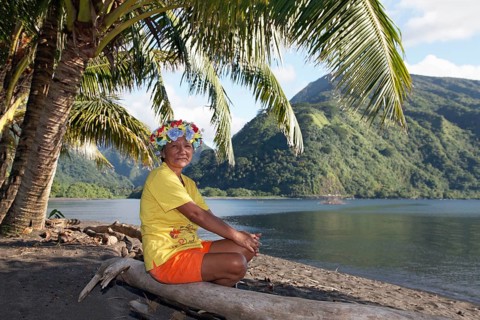 Old woman with wreath of flowers sitting under palm trees on tree trunk