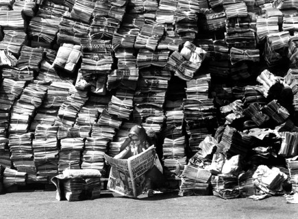 Woman reads newspaper in front of a big pile of newspapers
