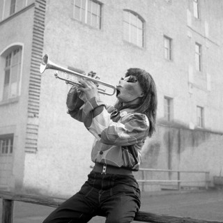 Man with shirt and mask sits on a wooden railing and plays trumpet