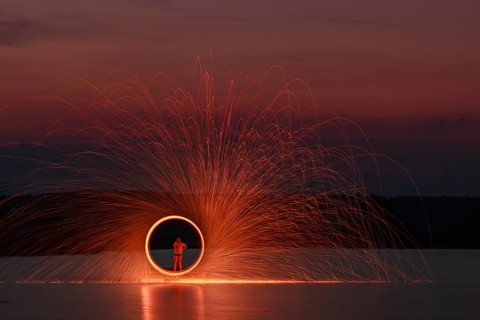 A man stands on a platform in a lake and creates a hoop of fire with glowing steel wool around him