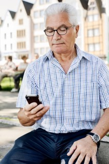 Grey-haired senior sits on a wall with his smartphone in his hand