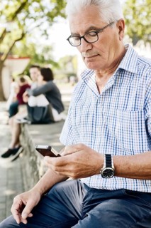 Grey-haired senior sits on a wall with his smartphone in his hand