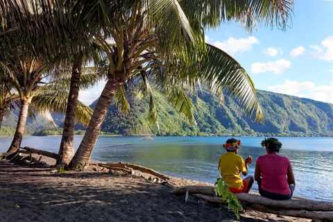 Two women with wreath of flowers sitting under palm trees on tree trunk