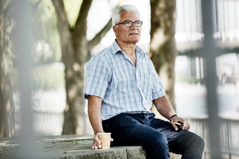 Senior sits with his Smartphone and a Coffe to go on a wall at Cologne's Rhine bank Promenade