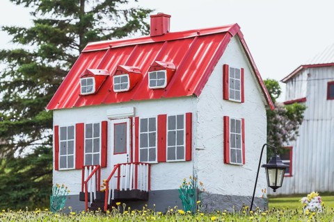 White with red trim miniature reproduction of an old Canadiana cottage style home facade in a field of yellow dandelion flowers in late spring