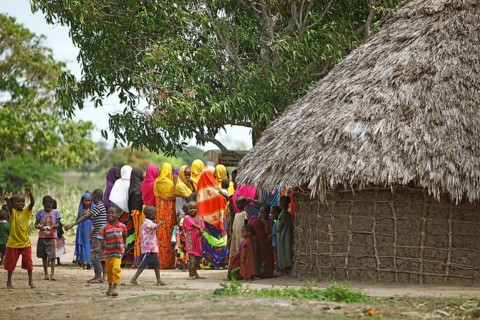 Women and children in colourful clothing next to mud hut