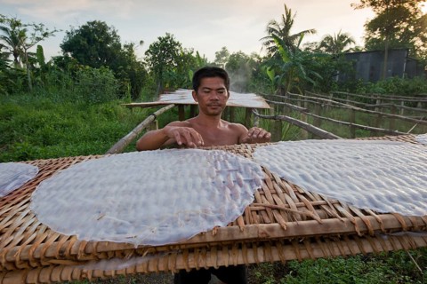 Vietnamese worker spreads steaming rice noodle patties on bamboo frame to dry