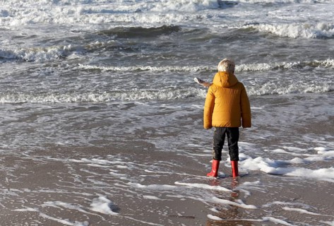 Little boy in winter clothes standing in the surf on the beach
