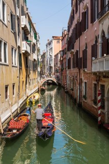 Gondolier steering gondola with tourists on narrow canal with historical buildings