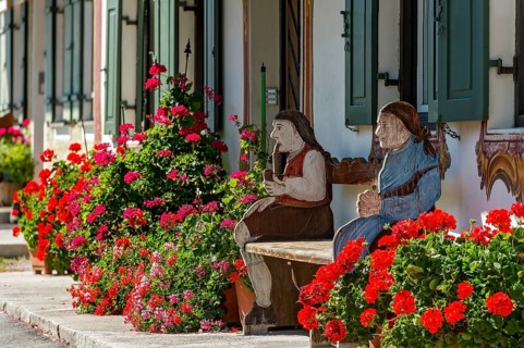 Geraniums (Pelargonium) and traditional Bavarian Bench with figures of farmer and his wife in front of farmhouse