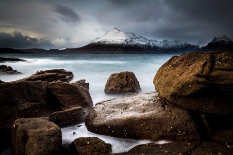 Big rocks in the water of the North Sea with snow-covered Cullin mountains in the background