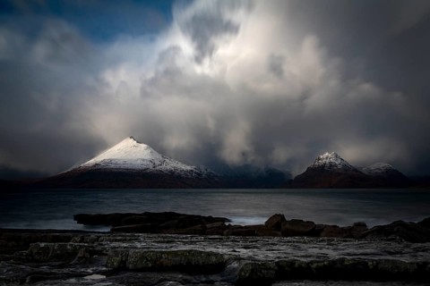 Big rocks in the water of the North Sea with snow-covered Cullin mountains in the background