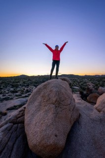 Young woman standing with outstretched arms on round granite rocks at sunset