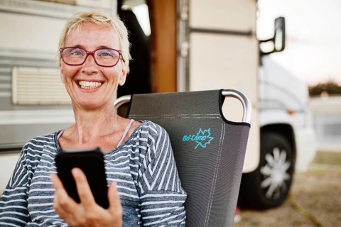 Woman sits with smartphone in a camping chair in front of her camper van