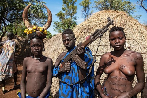 Young girls with decorative scars and man with Kalashnikov in front of hut