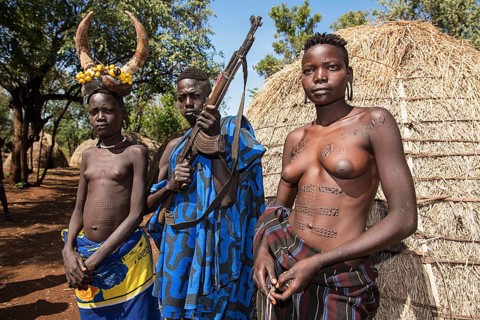 Young girls with decorative scars and man with Kalashnikov in front of hut