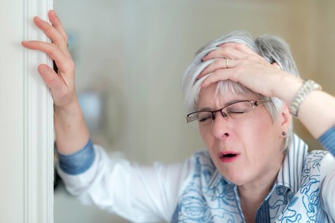 Senior woman with a headache holds her hand on her forehead and props herself on the door frame