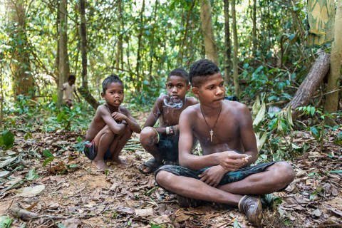 Three boys of the Orang Asil tribe sitting on the ground in the jungle and smoking