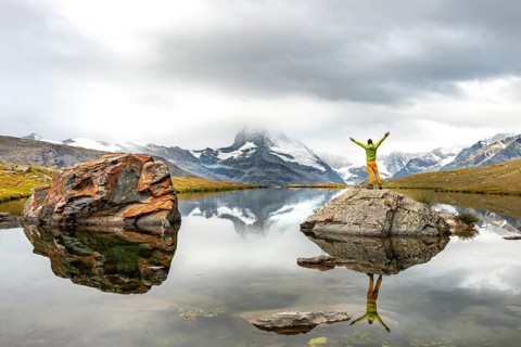Hiker stands on rocks in the lake and stretches his arms into the air