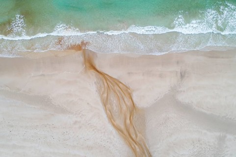 Creek with reddish ferruginous water flows over a sandy beach into the sea
