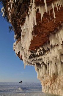 Icicles hanging from red sandstone cliff and people walking on frozen Lake Superior