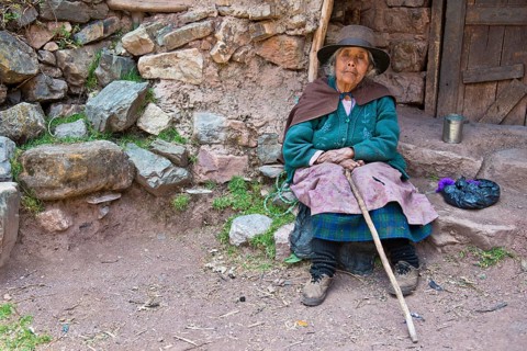 Mature Quechua Indian woman wearing a hat sitting on a stone in front of a doorway