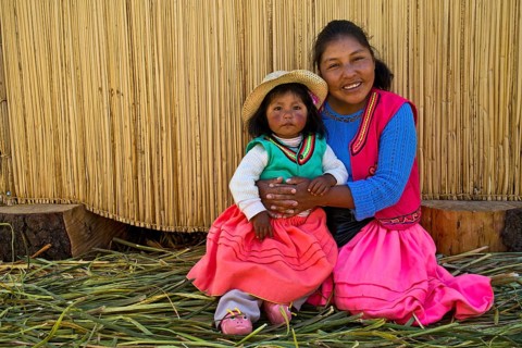 Friendly woman and a girl of the Uro Indians wearing traditional dress sitting in front of a reed hut