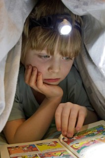Boy reading a comic with a head lamp under the duvet