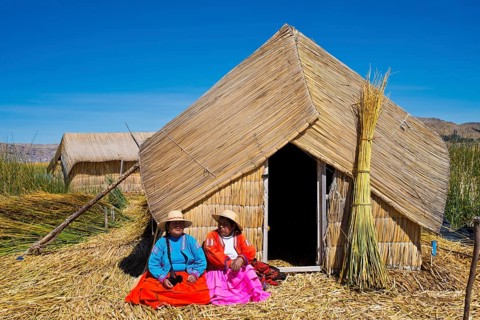Two women of the Uro Indians wearing traditional dress sitting in front of a reed hut
