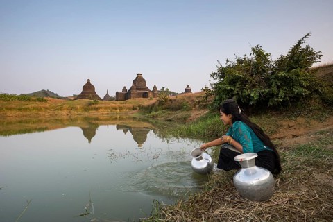 Smiling girl drawing water from a water hole with a vessel made of aluminium