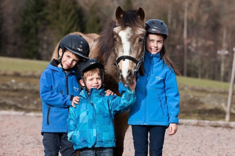 Three children wearing riding helmets standing beside a pony