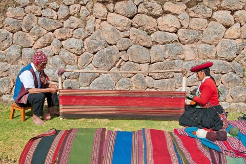 Two Quechua Indians in traditional dress squatting on the floor working on the stretcher of loom