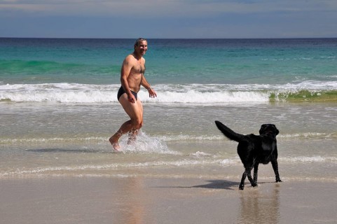 Man in swimming trunks playing with a dog on the beach of Rispond Bay