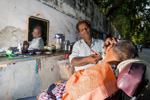 A barber is shaving a customer at his open-air shop