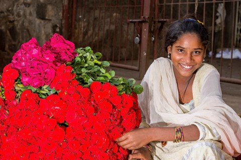 A smiling young woman in a white sari is selling red roses at the weekly market