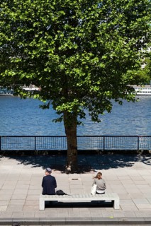 Two people sitting on concrete bench by river with tree