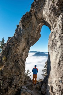 Young man stands in the rock gate Breitensteinfensterl with a view of the high fog above the valley
