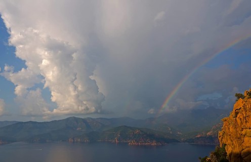 The typical bizarre red rocks of the Calanche of Piana and the mediterranean sea at the Gulf of Porto in the background below a rainbow and some clouds. The Calanche of Piana is in the western part of...